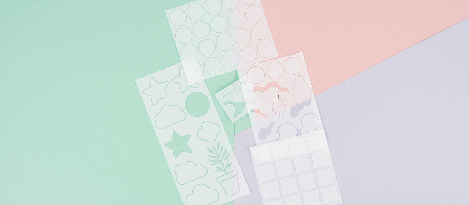 다양한 모양의 빈 비닐 스티커들이 파스텔 톤의 책상 위에 예술적으로 배열되어 있으며, 맞춤 디자인에 적합합니다.