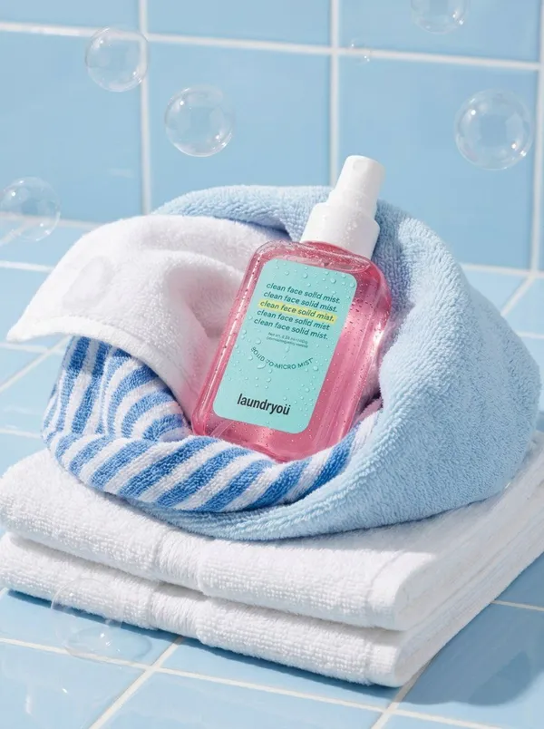 A clean commercial product photo of a translucent pink facial mist bottle on blue tiles and towels with bubbles.